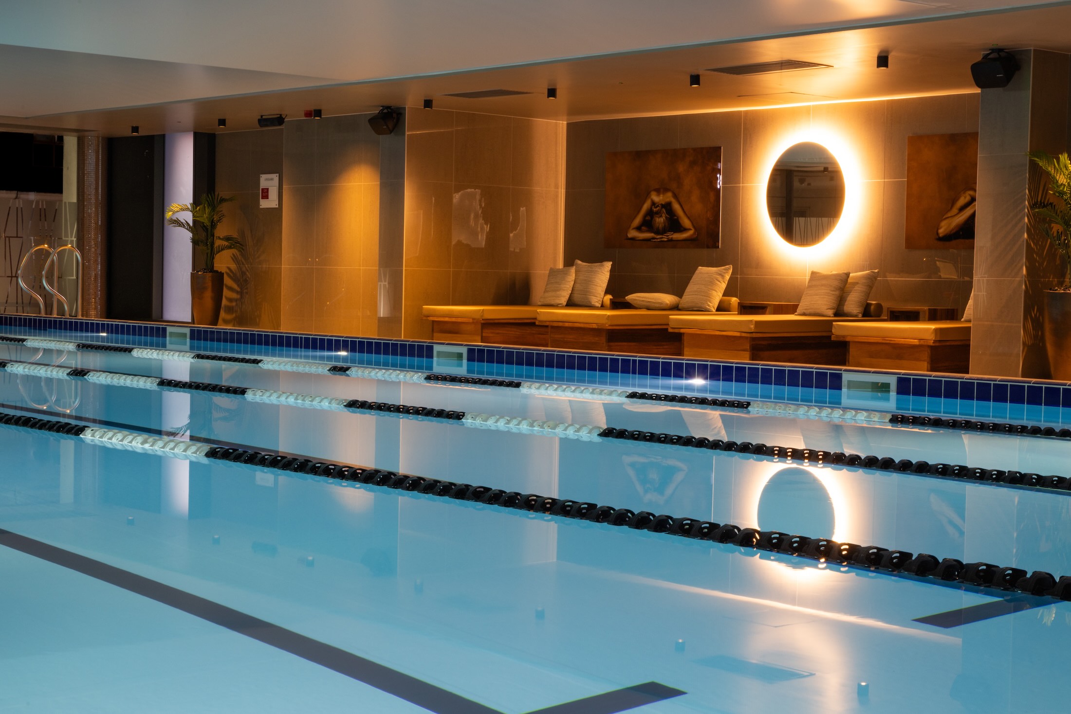 Indoor lap pool reflecting a warm spa lounge with cushioned daybeds and a glowing circular mirror.