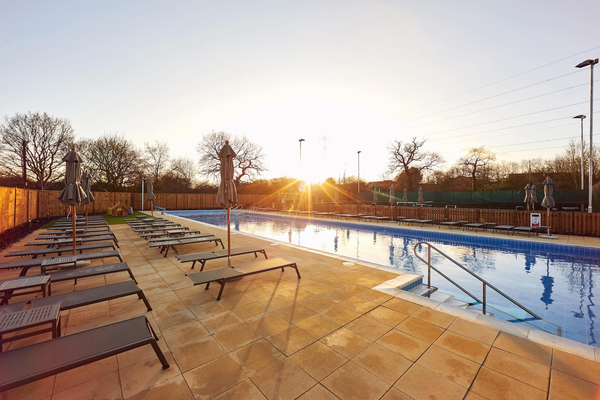 Outdoor swimming pool at sunset, lined with rows of empty lounge chairs on a tiled deck with sun umbrellas.