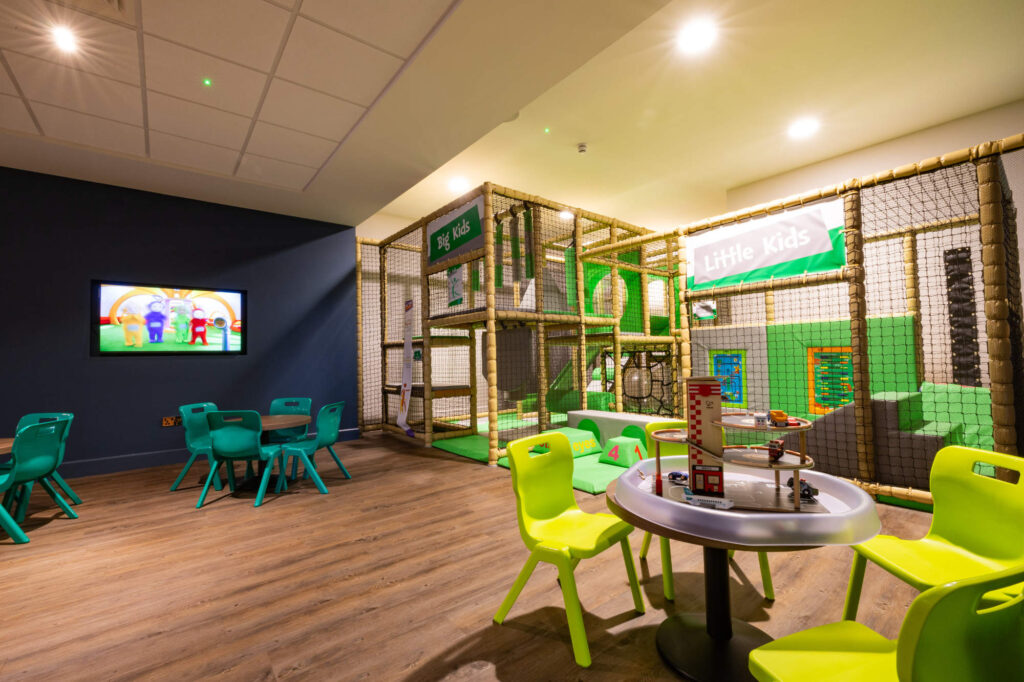 Indoor play area with a netted soft play structure, colorful child-sized tables and chairs, and a TV on the wall.