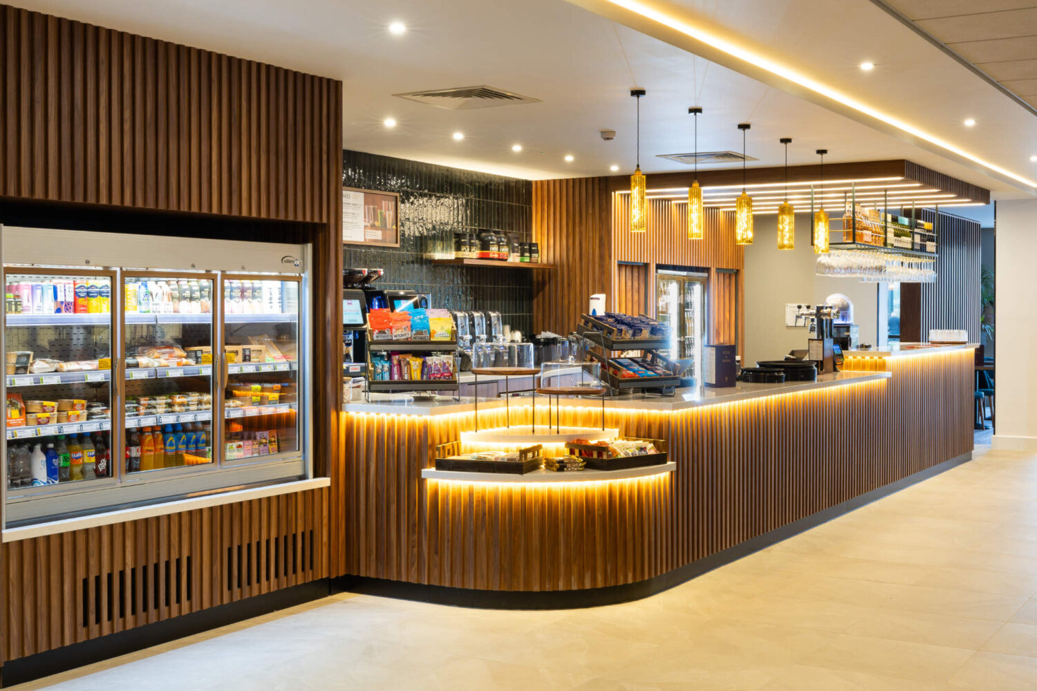 Modern cafe counter with vertical wood paneling, warm under-counter lighting, and a glass-door display fridge.