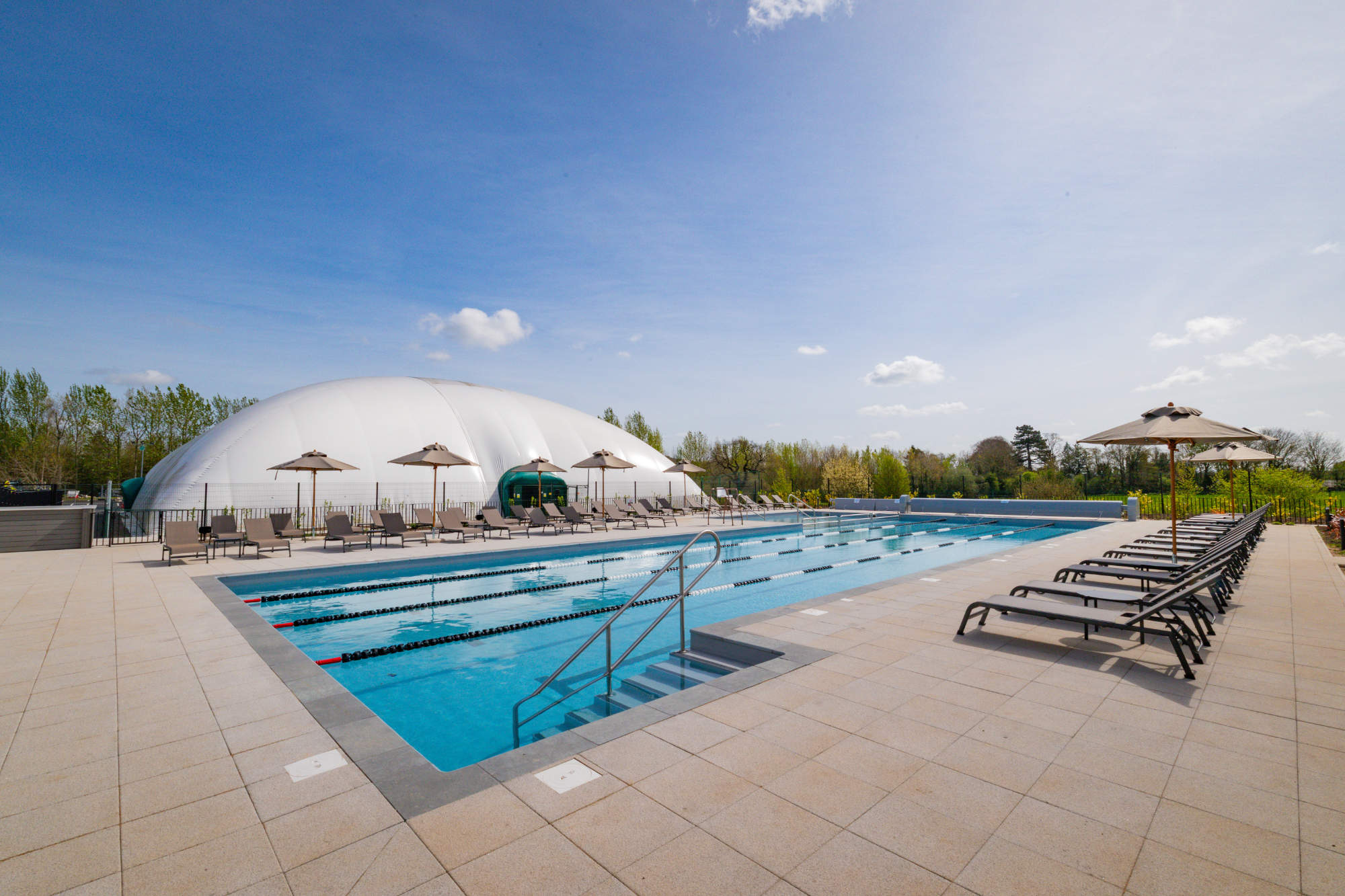 Outdoor lap pool lined with lounge chairs and umbrellas, with a large white dome structure in the background.