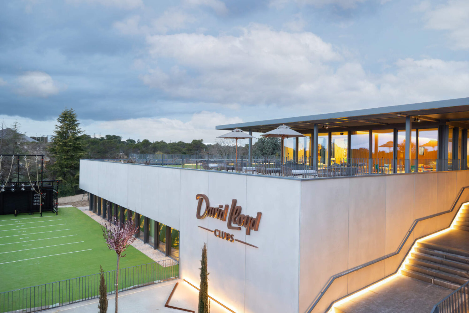 Modern David Lloyd Clubs facility featuring a rooftop terrace, illuminated stairs, and an outdoor turf field.
