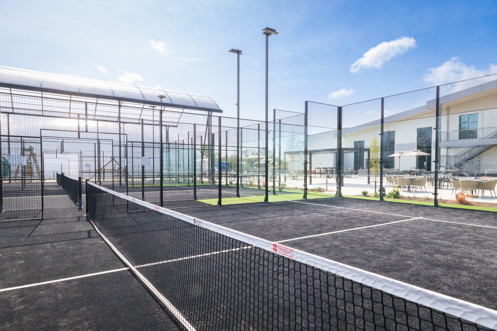 Outdoor padel tennis courts with a black surface, glass walls, and a net under a bright blue sky.