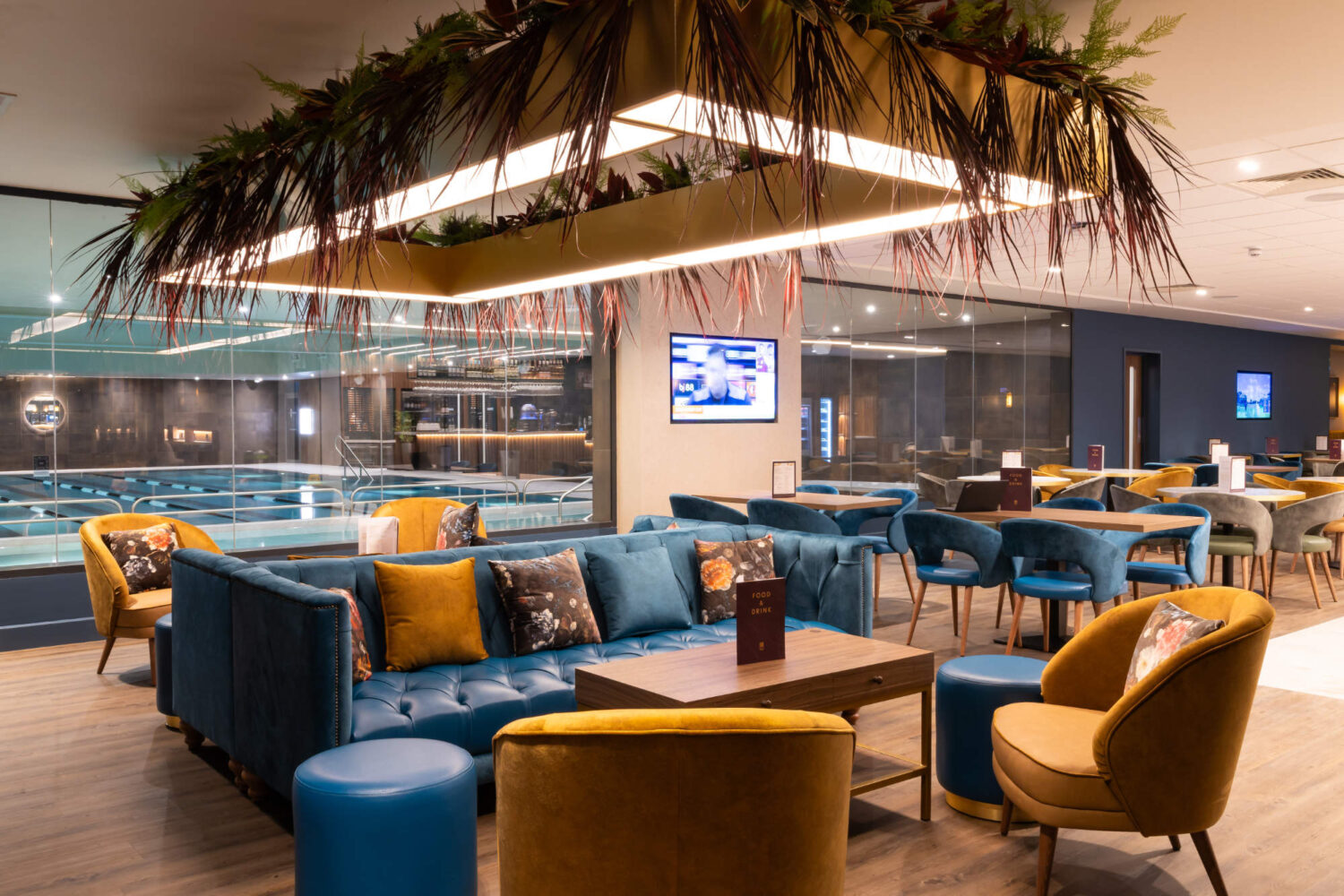 Modern lounge with blue and yellow seating, a hanging planter light, and an indoor pool visible through glass.